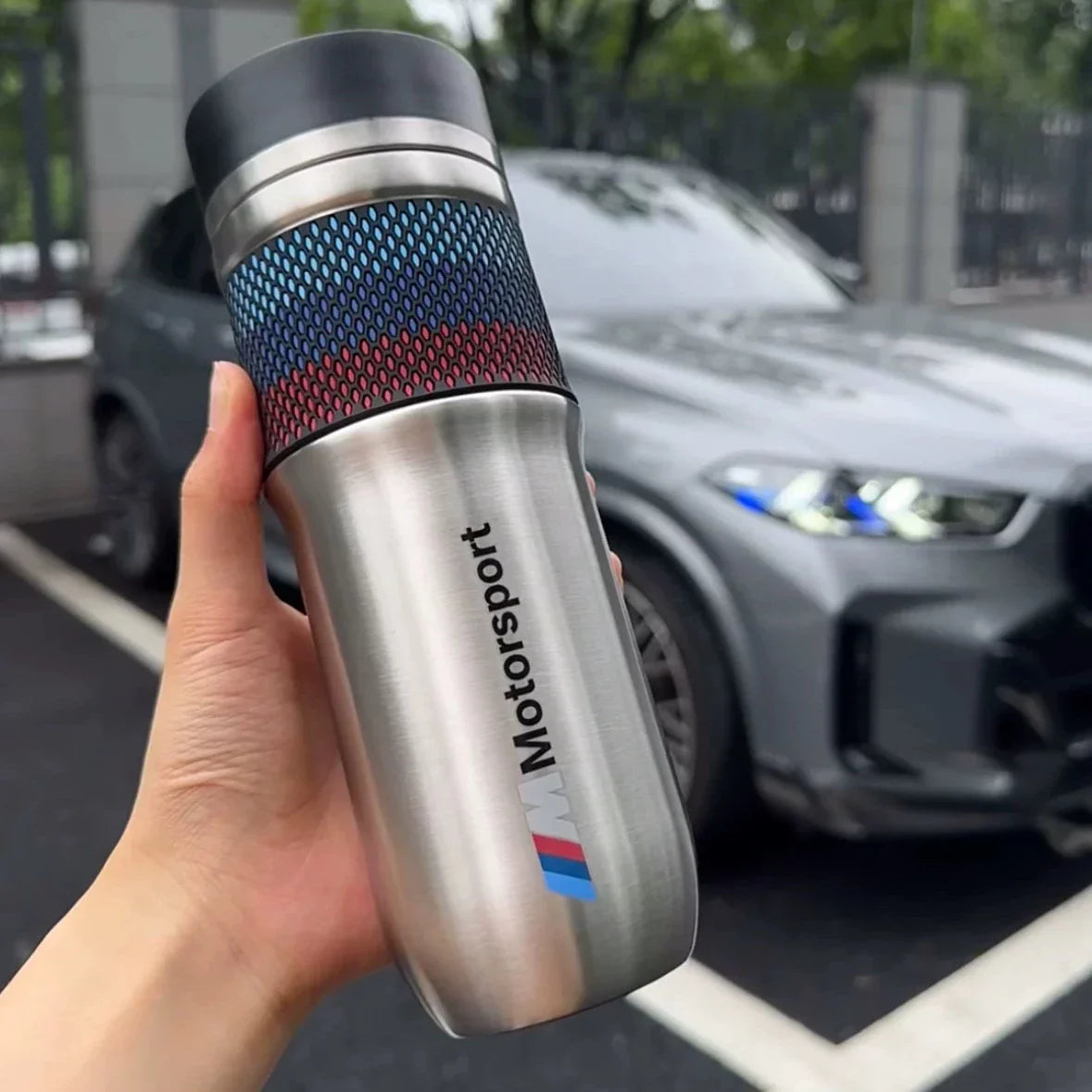 BMW M Motorsport stainless steel thermos cup held in hand with BMW car in background
